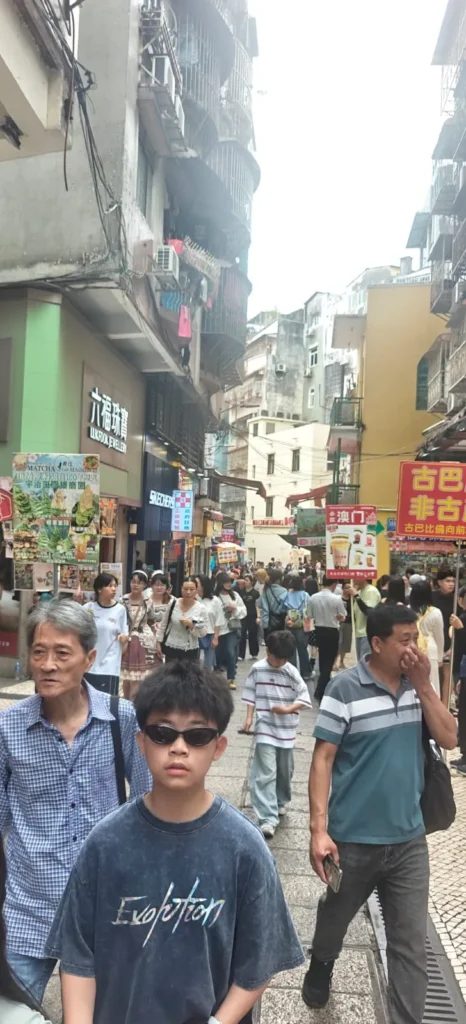 Una concurrida escena callejera en la ciudad de Shenzhen, con mucha gente paseando. Edificios con balcones se alinean a ambos lados de la calle, y coloridos carteles con texto en chino cuelgan sobre las tiendas. El cielo está parcialmente nublado.
