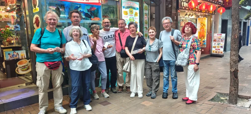 Un grupo de diez adultos de pie en una acera de la ciudad de Shenzhen, frente a restaurantes con letreros de colores y farolillos, algunos con comida o bebida en la mano, todos sonriendo a la cámara.