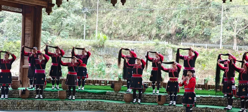 En Guilin, un grupo de artistas con trajes coordinados en negro y rojo, de pie sobre escalones de piedra al aire libre, sostienen largos objetos negros sobre sus cabezas, con vegetación y una estructura de madera de fondo.