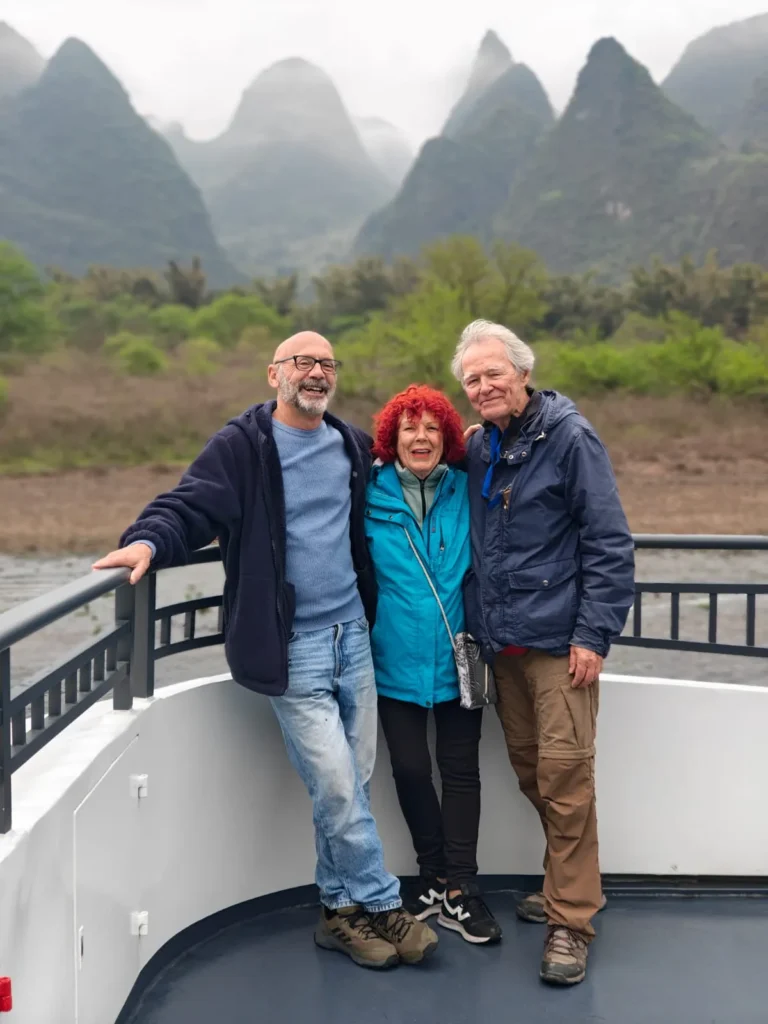 Tres personas permanecen juntas en la cubierta de un barco, sonriendo a la cámara. Detrás de ellos se ven las montañas de Guilin cubiertas de niebla y vegetación. El grupo va vestido con chaquetas informales y ropa de exterior.