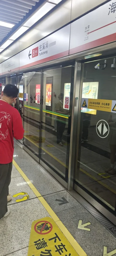 Un andén de metro con puertas de seguridad de cristal, carteles en chino e inglés y una persona con camisa roja esperando, típico de las estaciones modernas de Shenzhen. Aparecen reflejos y otros pasajeros en el lado opuesto del andén.