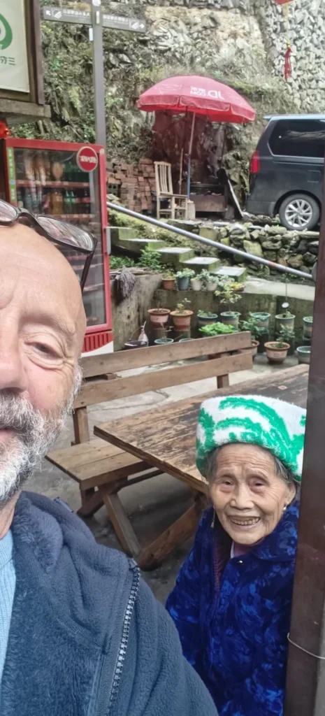 Un hombre y una anciana sonríen para hacerse un selfie al aire libre cerca de bancos de madera y macetas en Shenzhen. Al fondo se ven un frigorífico rojo, un coche negro y rocas.
