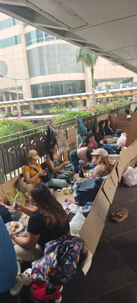 Un grupo de personas se sientan en el suelo al aire libre en Shenzhen, algunas sobre cajas de cartón desplegadas, comiendo y hablando. Hay bolsas y envases de comida desperdigados. Al fondo se ve un edificio moderno y vegetación.