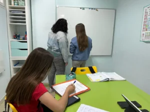 Tres chicas están en un aula de la Academia Pentalfa de Rivas. Dos están de pie junto a una pizarra, mientras que una está sentada en una mesa verde escribiendo en un cuaderno, inmersa en un Intensivo de PAU. Sobre la mesa hay libros abiertos, un estuche y un teléfono.