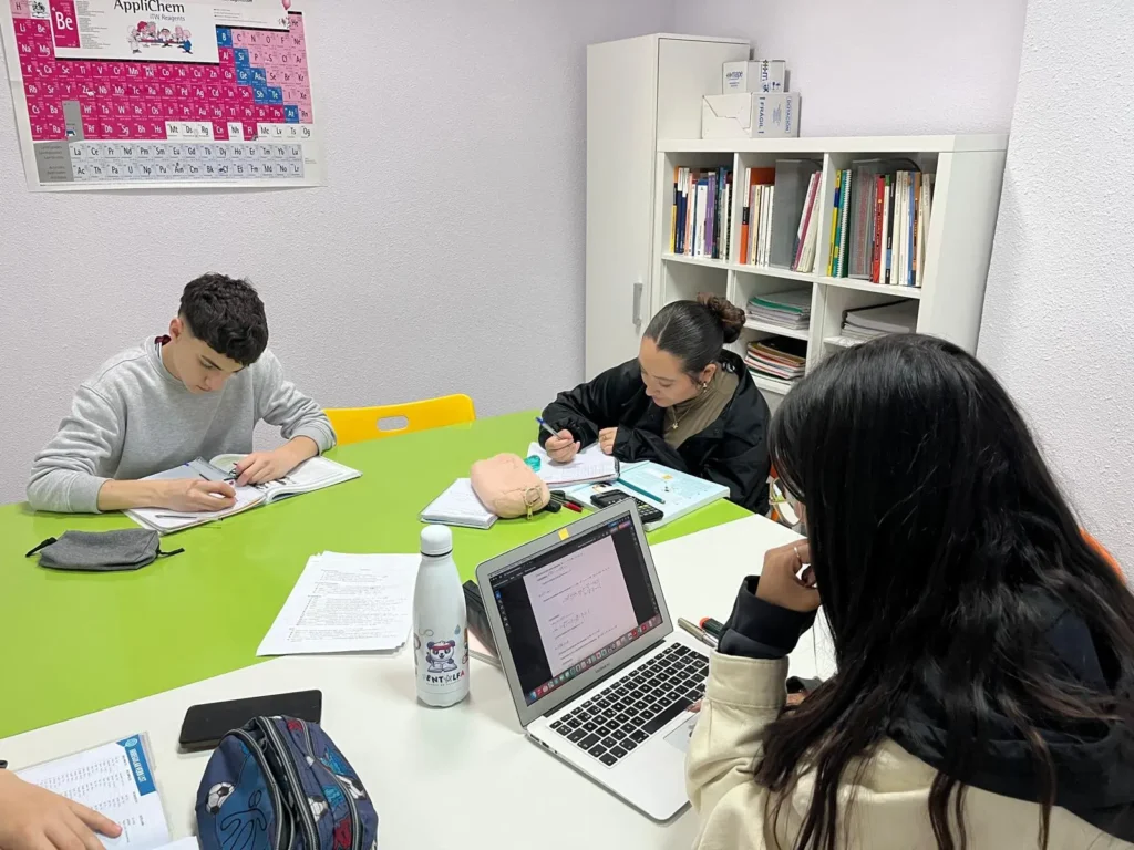 Tres estudiantes se sientan a estudiar alrededor de una mesa verde en la Academia Pentalfa. Uno escribe en un cuaderno, otro lee y el tercero trabaja en un portátil, preparándose para el Intensivo de PAU. Libros, papeles y estuches cubren la mesa; detrás, estanterías y una tabla periódica.