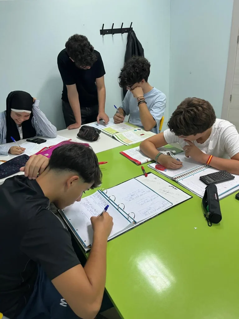 Cinco alumnos de la Academia Pentalfa de Rivas se sientan alrededor de una mesa verde con cuadernos, bolígrafos y una calculadora, preparando la PAU. Cuatro escriben mientras uno, de pie, repasa los apuntes. Las chaquetas cuelgan de un perchero al fondo.