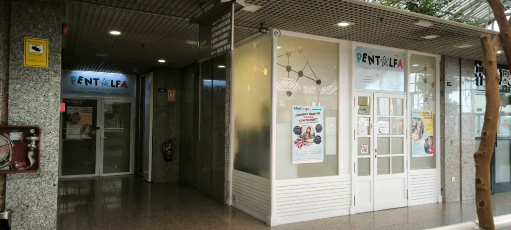 Un pasillo de Rivas conduce a la entrada acristalada de la Academia Pentalfa, con "PENTALFA" en la puerta de la oficina y carteles que promocionan su curso intensivo de PAU. La zona, bien iluminada, también cuenta con un extintor cerca.