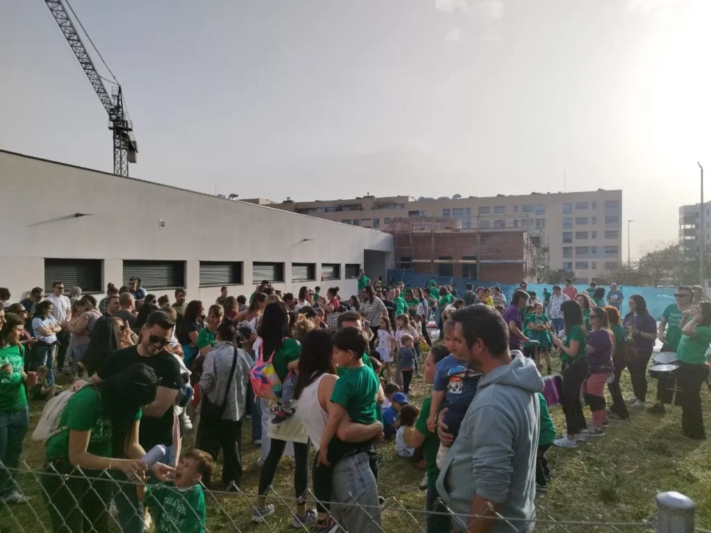 Un gran grupo de personas, entre adultos y niños, se reúnen al aire libre en una zona de césped cerca del CEIP Mercedes Vera. Muchos llevan camisetas verdes. Una grúa y varios edificios de apartamentos señalan obras en curso en el fondo.
