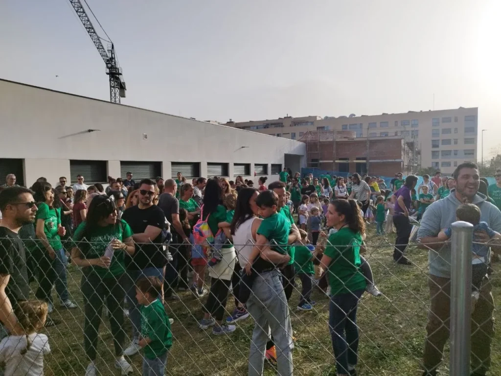 Un nutrido grupo de personas, muchas de ellas con camisetas verdes, se reúnen al aire libre cerca del CEIP Mercedes Vera, en una zona de césped detrás de una valla metálica. Algunos adultos tienen niños en brazos y charlan, mientras al fondo se ven obras y edificios de apartamentos con una grúa.