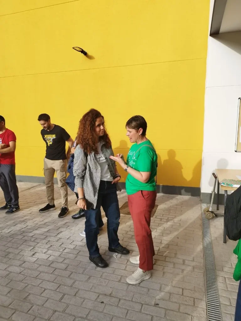 Dos mujeres conversan en el CEIP Mercedes Vera, cerca de una pared amarilla brillante, mientras otras tres están cerca; una mujer gesticula con la mano mientras conversan a la luz del sol.