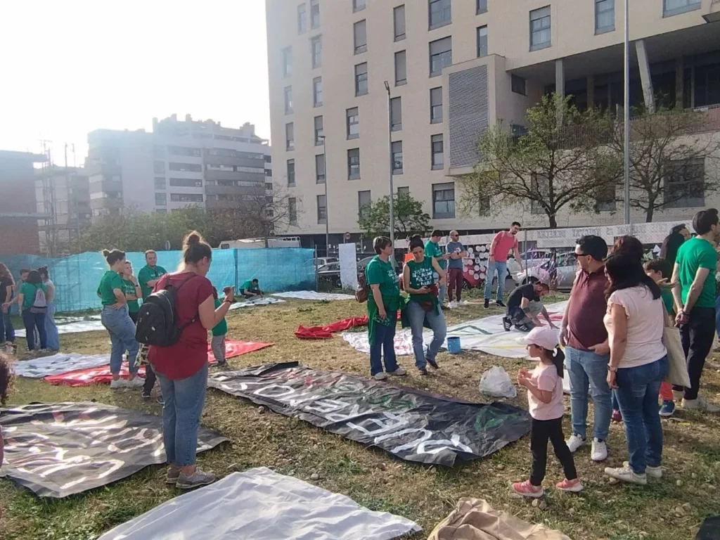 Un grupo de personas, entre las que hay niños y adultos, conversan cerca de un edificio junto a pancartas y sábanas colocadas sobre el césped. Algunos llevan camisetas verdes mientras se reúnen en el CEIP Mercedes Vera para hablar de obras, con edificios urbanos al fondo.