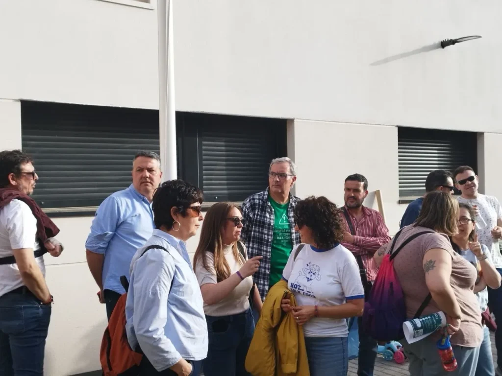 Un grupo de personas se encuentra frente al CEIP Mercedes Vera, un edificio blanco con persianas negras cerradas. Algunos hablan, llevan bolsos o chaquetas, y uno de ellos lleva un tatuaje en el brazo. El grupo parece estar reunido casualmente durante el encierro.