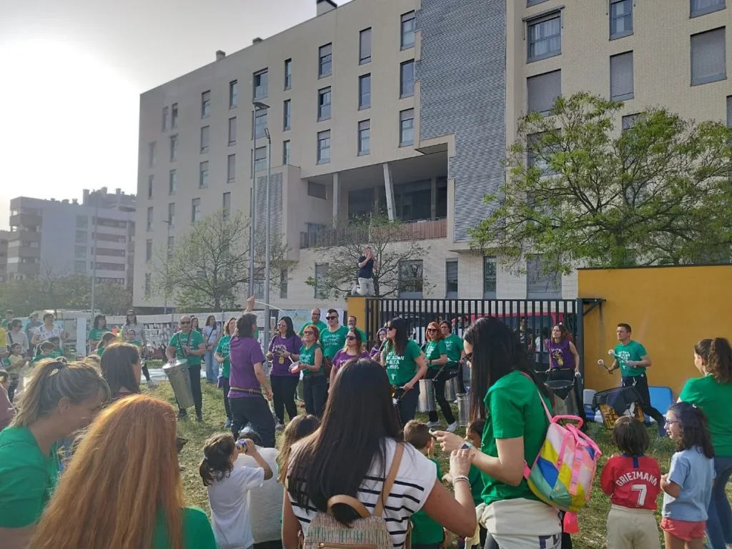 Un grupo de personas, muchas con camisetas verdes, se reúnen al aire libre cerca del CEIP Mercedes Vera, un edificio moderno. Algunos tocan instrumentos musicales mientras otros observan. El cielo está despejado y entre la multitud hay varios niños.
