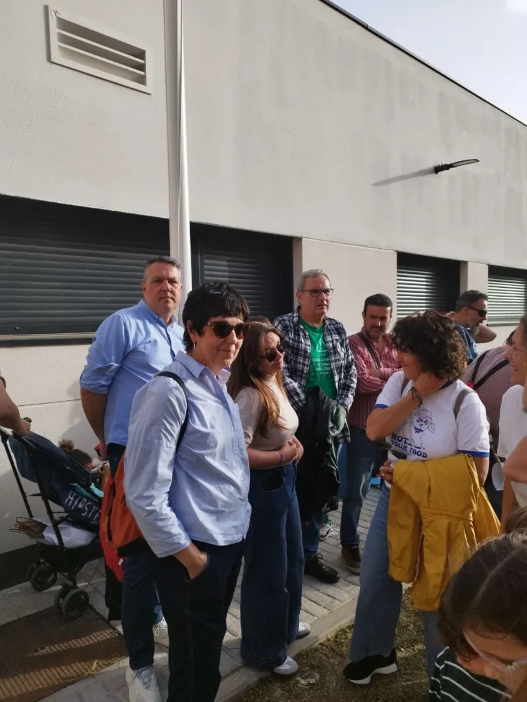 Un grupo de personas se encuentra frente al CEIP Mercedes Vera, un edificio de paredes blancas y ventanas enrejadas. Algunos hablan y visten ropa informal. A la izquierda se ve un cochecito, posiblemente debido al encierro de las obras en curso.