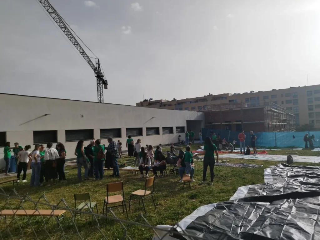 Un grupo de personas se reúne cerca del CEIP Mercedes Vera, en una zona de césped con sillas y lonas esparcidas. Al fondo se ve una grúa de obras cercanas y edificios de viviendas más alejados.