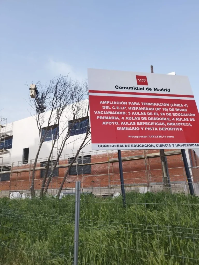 Una obra con un gran cartel blanco y rojo en español que detalla los planes de ampliación del CEIP Mercedes Vera en Madrid, incluidas nuevas aulas e instalaciones. Detrás del cartel se ve un edificio parcialmente construido y una valla.
