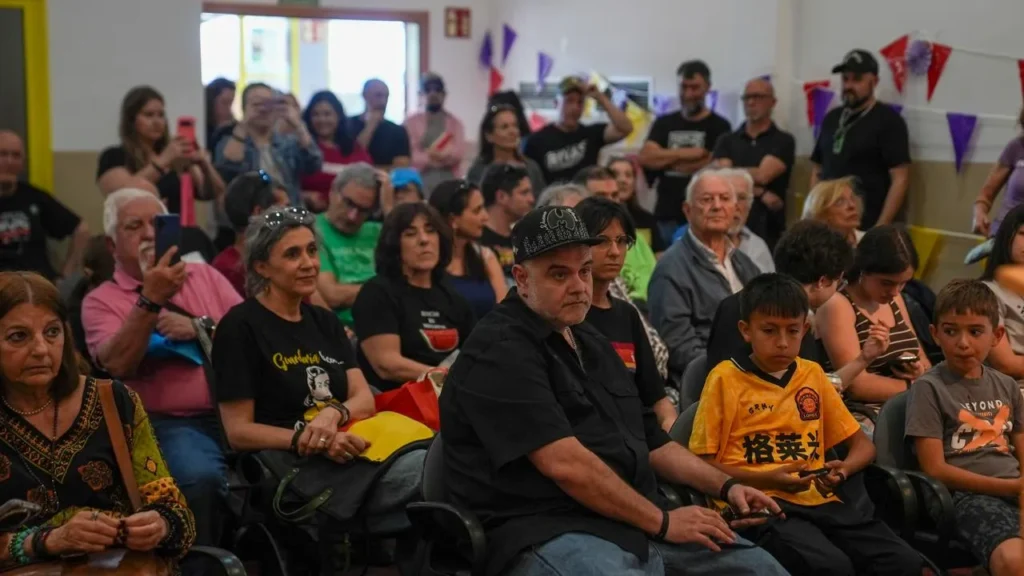 Un nutrido grupo de personas, entre las que hay adultos y niños, se sientan muy juntas en un espacio interior muy iluminado de las Jornadas Republicanas de Rivas. Algunos llevan ropa informal y sombreros. En las paredes cuelgan banderas y adornos de colores, y hay cerca de 500 personas presentes.