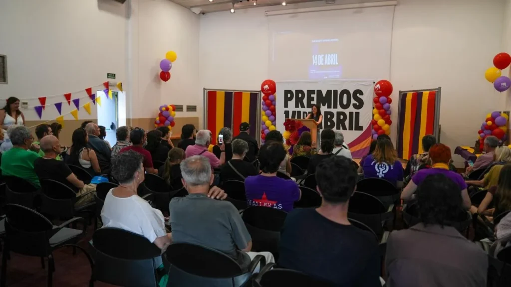 Un grupo de personas se sienta en un salón de actos de Rivas, frente a un escenario decorado con globos de colores y pancartas de los Premios Abril. Un orador, de pie, se dirige al público durante las Jornadas Republicanas. En la pared del fondo se ve una pantalla.