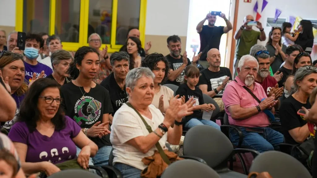 Un grupo de personas de distintas edades permanecen sentadas muy juntas en una sala muy iluminada de la Casa de Asociaciones, algunas aplaudiendo y otras, de pie, haciéndose fotos al fondo.
