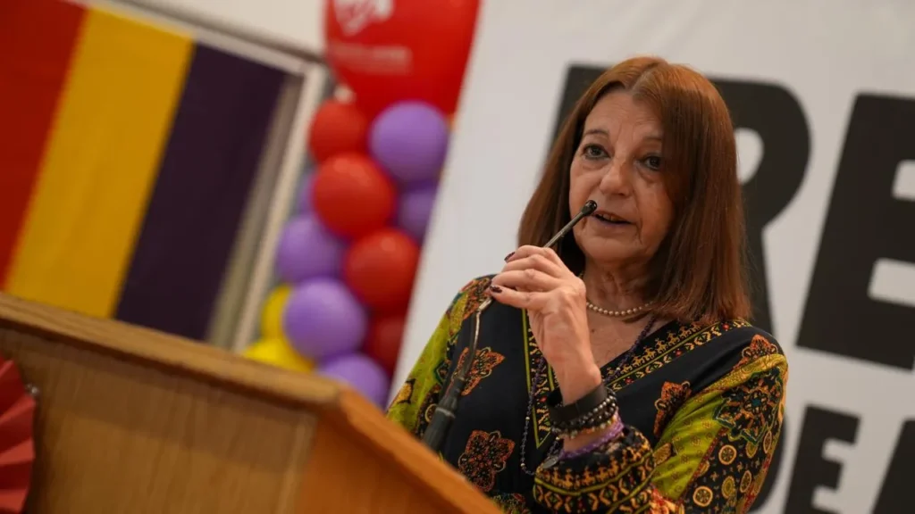 Una mujer de pelo castaño liso, de pie en un estrado, se lleva un bolígrafo a la boca mientras se dirige al público. Al fondo se ven parcialmente globos de colores y una pancarta.