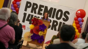 Una mujer sube a un podio decorado en Rivas, dirigiéndose a 500 personas durante las Jornadas Republicanas, con globos de colores y adornos de papel enmarcando una gran pancarta de PREMIOS 14 DE ABRIL detrás de ella.
