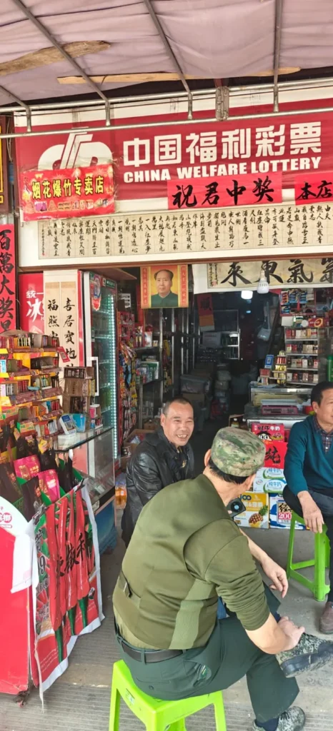 Tres hombres conversan sentados en taburetes verdes frente a una pequeña tienda de Shenzhen bajo un cartel que reza "Lotería del Bienestar de China". En las estanterías de la tienda hay aperitivos, bebidas y billetes de lotería, mientras pancartas y carteles rojos decoran la entrada y las paredes.