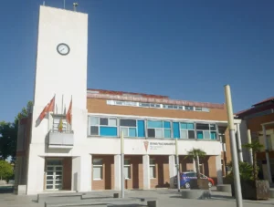 Un edificio de dos plantas del Ayuntamiento de Rivas con una torre del reloj, varias banderas y un cartel en el que se lee Rivas Vaciamadrid Alcaldía; un coche de policía está aparcado fuera bajo un cielo azul despejado.
