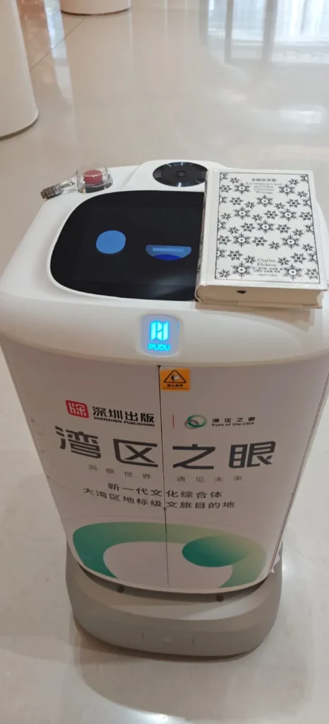 Un robot de servicio blanco con una pantalla digital en la cara se encuentra en un interior de Guilin. Tiene texto y logotipos chinos en la parte frontal, además de un cuaderno estampado colocado en la parte superior cerca de un sensor y un botón de parada de emergencia.