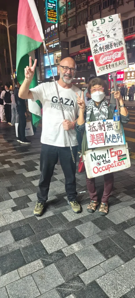 Dos personas de pie en una calle nocturna de Macao, con carteles a favor de Palestina y en contra de la ocupación. Una ondea una bandera y lleva una camiseta de GAZA PALESTINA; la otra lleva una máscara. Unos letreros luminosos iluminan el fondo.