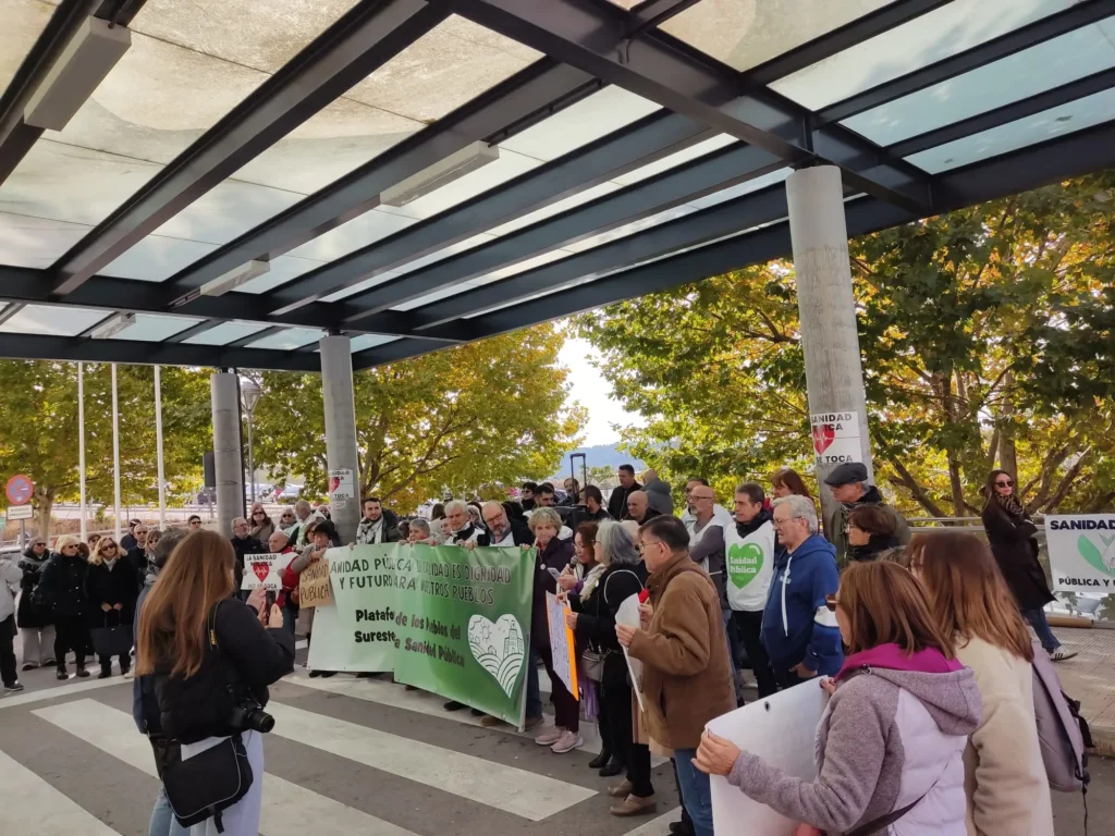 Un grupo de personas protesta bajo un toldo de cristal con pancartas y carteles sobre Sanidad Pública y la falta de personal en el Hospital del Sureste. Algunos hacen fotos y se ven árboles al fondo.