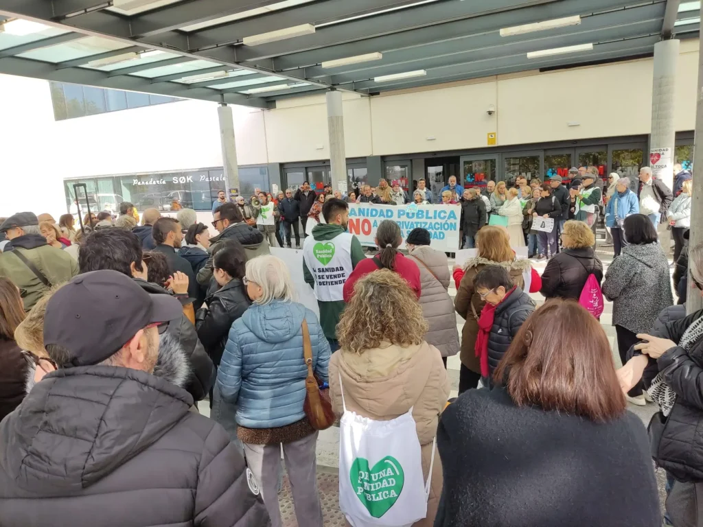Una multitud se reúne al aire libre cerca de la entrada de un edificio, muchos con pancartas y carteles. Una de las pancartas más destacadas habla de la defensa de la sanidad pública y de la oposición a la privatización del Hospital del Sureste, destacando la preocupación por la falta de personal.