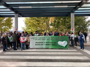 Un grupo de personas en un paso de peatones con una pancarta que dice Sanidad pública de calidad es dignidad y futuro para nuestros pueblos en una manifestación pública, exigiendo soluciones a la falta de personal en el Hospital del Sureste, con árboles cerca.