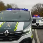 Un furgón de la Guardia Civil de Tráfico y un coche patrulla con luces intermitentes están parados en una carretera, cerca de conos naranjas y agentes de policía, junto a un vehículo de emergencias amarillo. Borrador automático se anota como parte del informe del incidente. Al fondo, árboles otoñales.