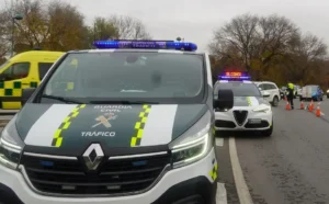 Un furgón de la Guardia Civil de Tráfico y un coche patrulla con luces intermitentes están parados en una carretera, cerca de conos naranjas y agentes de policía, junto a un vehículo de emergencias amarillo. Borrador automático se anota como parte del informe del incidente. Al fondo, árboles otoñales.
