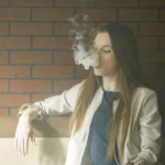 Una persona con el pelo largo se sienta en un sofá contra una pared de ladrillo, exhalando una nube de vapor mientras sostiene un vapeador, reflejo del auge del vapeo adolescente en España.