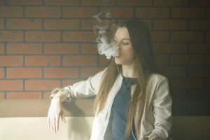 Una persona con el pelo largo se sienta en un sofá contra una pared de ladrillo, exhalando una nube de vapor mientras sostiene un vapeador, reflejo del auge del vapeo adolescente en España.