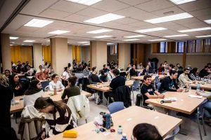 En una sala bien iluminada, un grupo numeroso de personas se sienta a la mesa para hablar y conversar. En las mesas se ven botellas, aperitivos y objetos personales. La sala tiene paredes de color beige e iluminación fluorescente.