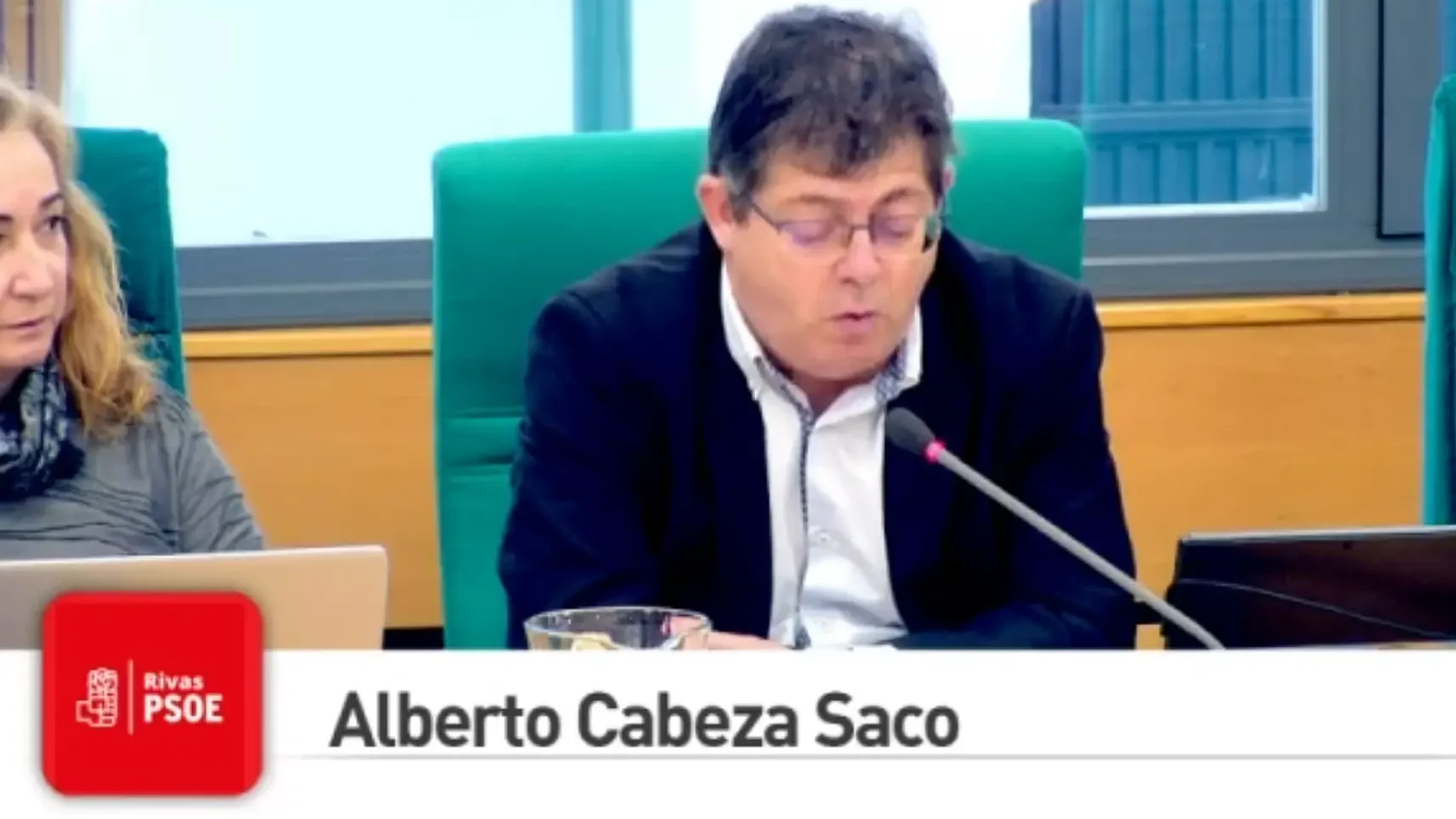 Un hombre con gafas y pelo corto y oscuro habla por un micrófono desde un escritorio, con una mujer a su lado. El texto superpuesto dice Alberto Cabeza Saco e incluye el logotipo del PSOE de Rivas en la esquina inferior izquierda.