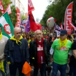 Un grupo de personas se sitúa al frente de una gran protesta callejera en Rivas, con camisetas de UGT Madrid y pañuelos amarillos, con coloridas banderas y pancartas de fondo. La multitud llena la calle detrás de ellos.