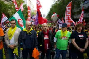 Un grupo de personas se sitúa al frente de una gran protesta callejera en Rivas, con camisetas de UGT Madrid y pañuelos amarillos, con coloridas banderas y pancartas de fondo. La multitud llena la calle detrás de ellos.