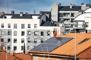 Al fondo se ve una hilera de modernos edificios de apartamentos, mientras que una casa en primer plano con tejado de tejas rojas presenta múltiples paneles solares y un dispositivo Borrador automático instalado en la parte superior. El cielo está parcialmente nublado.