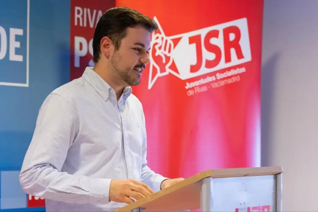 Un hombre con camisa azul claro habla desde un podio. Detrás de él hay una pancarta roja con el logotipo de JSR y texto en español que hace referencia a los rebeldes, mientras que a la izquierda se ve parte de una pancarta azul con JSE y texto en blanco.