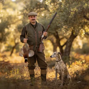 Un hombre vestido de cazador y con gorra plana se encuentra al aire libre sosteniendo una escopeta y un faisán, con su fiel perro a su lado entre árboles iluminados por el sol, capturando el espíritu del campo en medio de debates como la Manifestación contra la Ley de Caza.