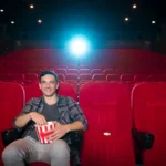 Un hombre se sienta solo en una sala de cine casi vacía, disfrutando de las palomitas en una butaca roja bajo el brillante proyector, aprovechando la oferta de Cine Senior para mayores, entradas a sólo 2 euros.