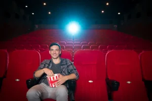 Un hombre se sienta solo en una sala de cine casi vacía, disfrutando de las palomitas en una butaca roja bajo el brillante proyector, aprovechando la oferta de Cine Senior para mayores, entradas a sólo 2 euros.