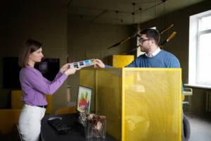 Dos personas en una oficina, probablemente de una agencia de marketing, examinan juntas una muestra de color; una mujer sujeta un extremo y un hombre se sienta en un escritorio sujetando el otro, con un ordenador y un separador amarillo entre ellos.