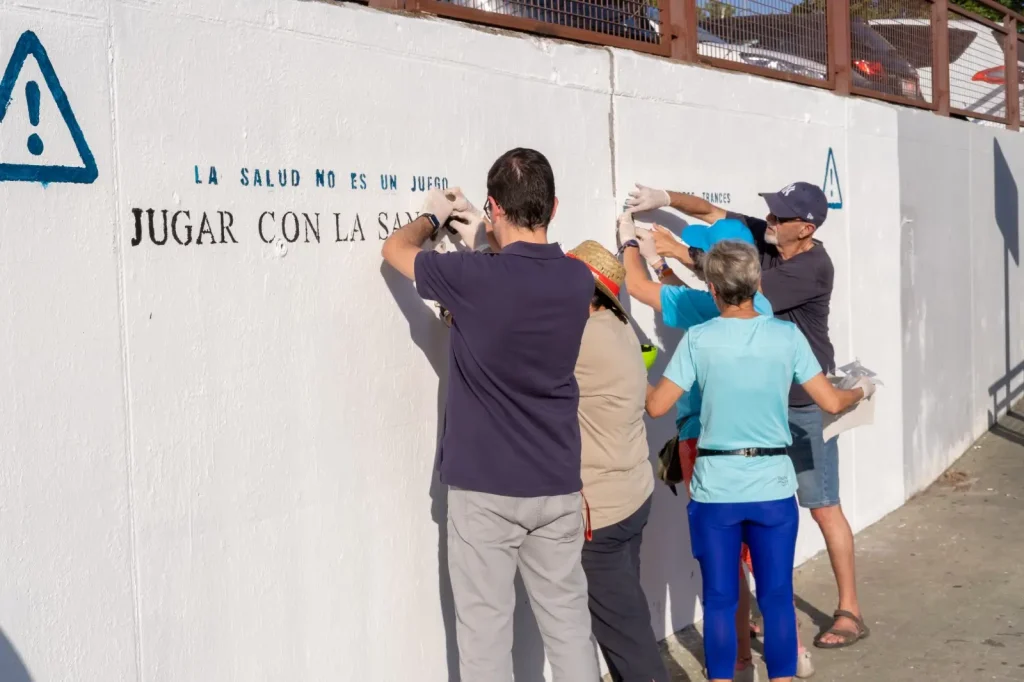 Un grupo de personas permanece junto a una pared blanca de Rivas, pintando o estarcido con letras negras y azules que forman un mensaje en español sobre la sanidad pública. Algunos llevan guantes y gorros. La escena tiene lugar al aire libre, a la luz del día.