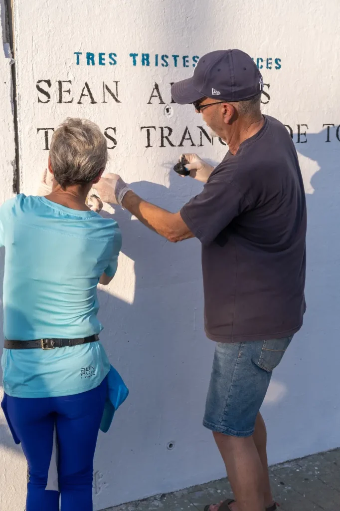 Dos personas vestidas de azul pintan letras negras -quizá poesías- en una pared blanca. Una de ellas está de cara a la pared, mientras que la otra, con gorra, sostiene pintura y un pincel. La luz del sol proyecta sombras sobre esta escena creativa, que recuerda el espíritu de Rivas.