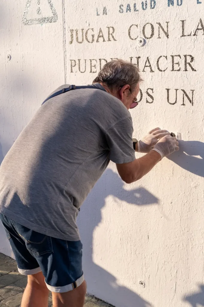 Un hombre mayor con camiseta gris, pantalones cortos azules y guantes pinta letras negras en una pared blanca exterior. La luz del sol proyecta su sombra, y algunas poesías españolas son parcialmente visibles en la pared.