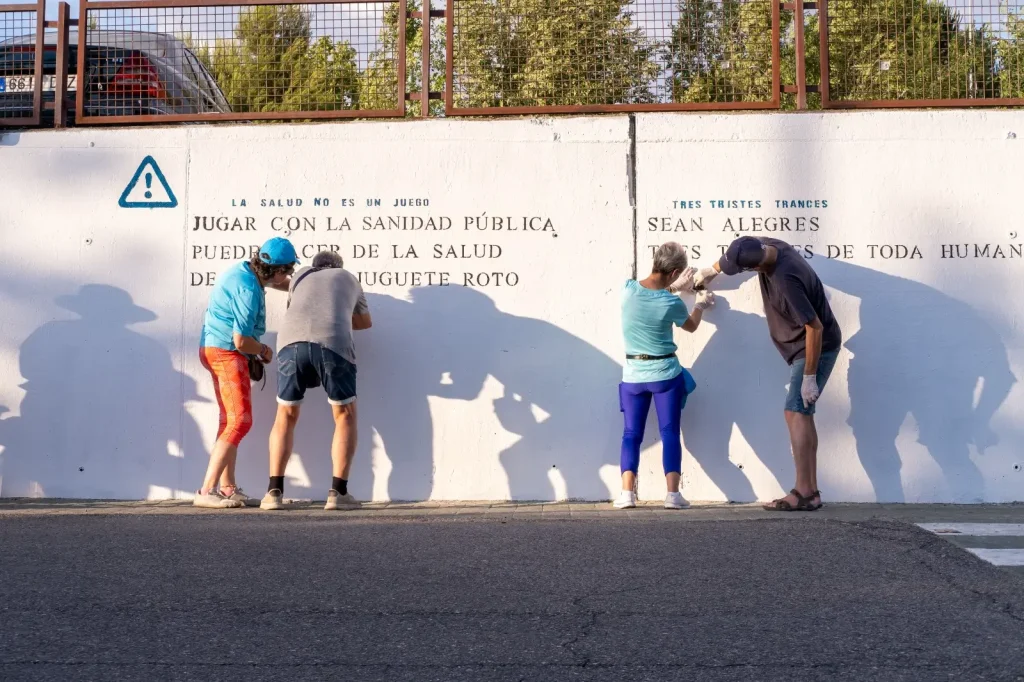 Cuatro personas permanecen de pie junto a una pared blanca con texto en español, posiblemente poesías, proyectando largas sombras mientras escriben o examinan la pared. La escena se desarrolla al aire libre en Rivas, con árboles y una valla al fondo.